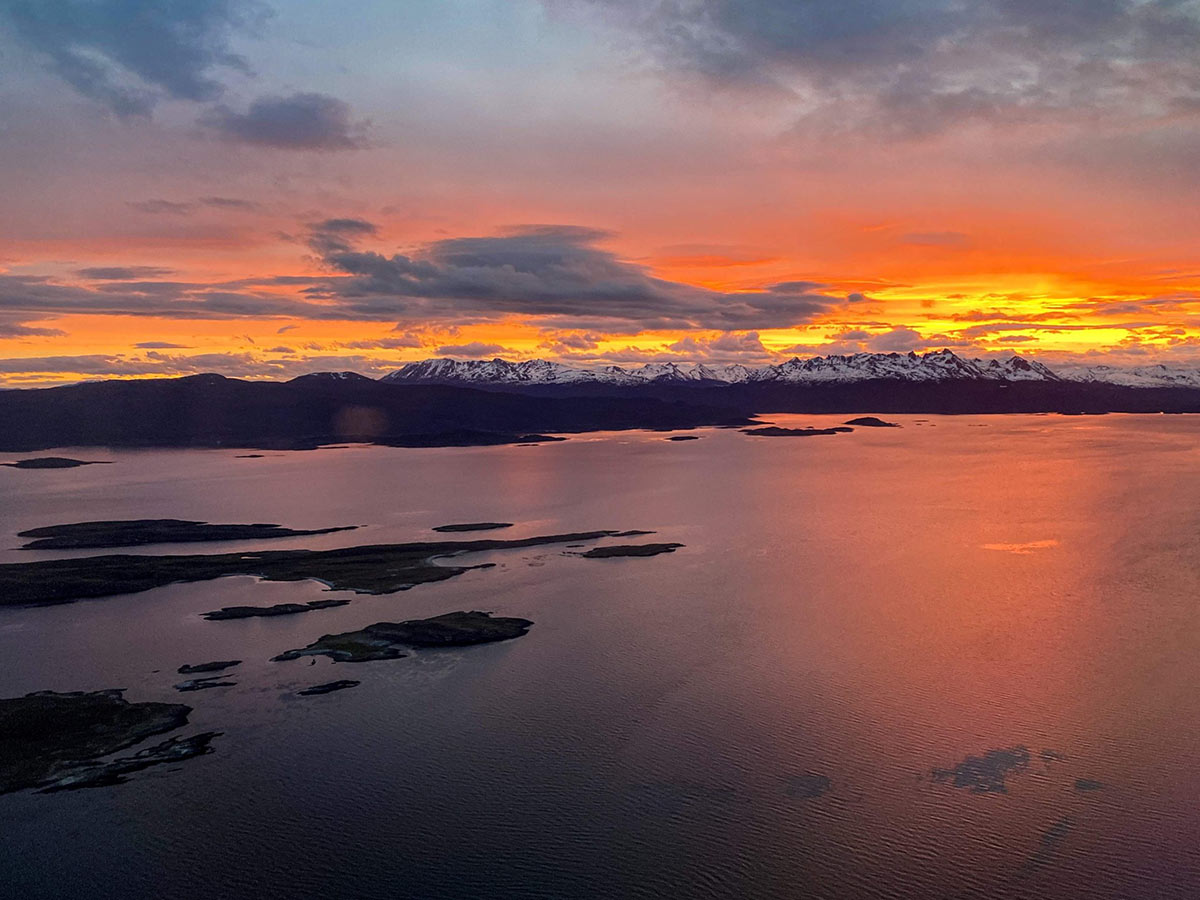 The mountains of Argentina with an orange and yellow sunrise or sunset behind them. There is a lake. There is snow on top of the mountains