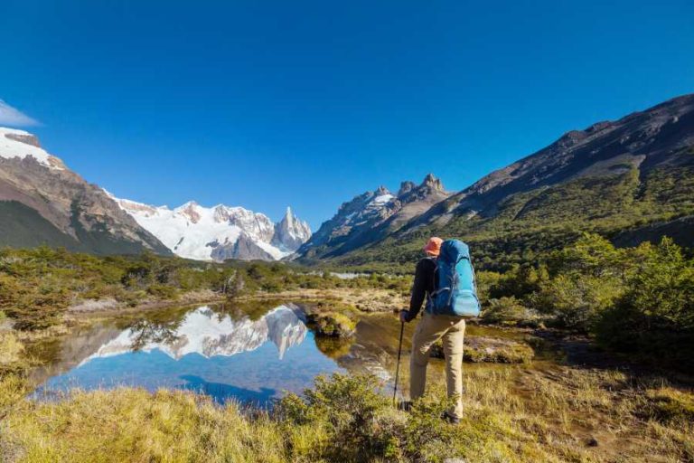 A hiker stands looking over a lake in the mountains of Patagonia. There are mountains surrounding the person and the blue sky reflects in the lake.